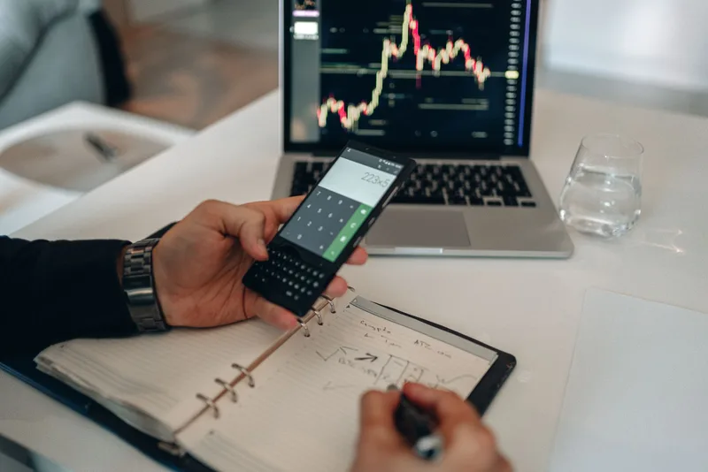 Hand holding a phone with a calculator open above a notebook of handwritten sums, with a laptop showing a price chart behind