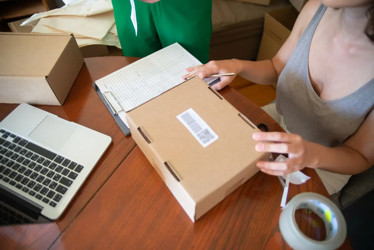 Woman at a wooden table closing a cardboard box with a barcode label, with a clipboard, laptop and roll of tape beside it