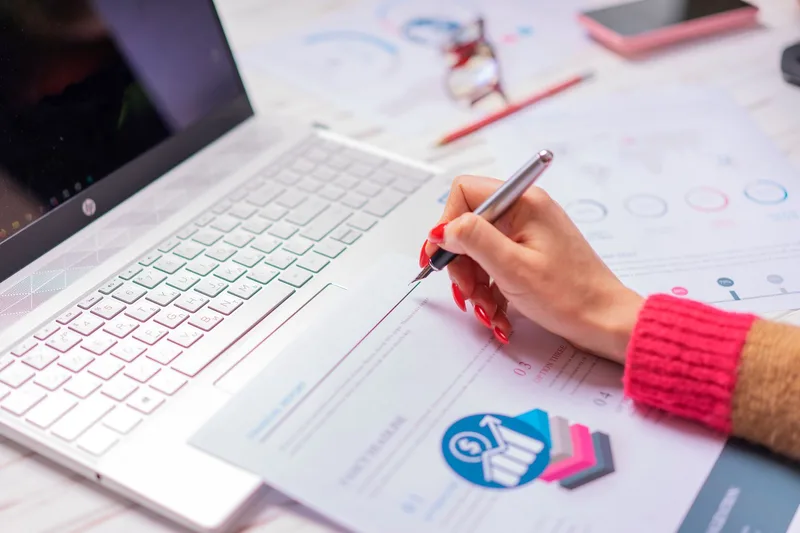 Person writing on paper beside a laptop while working through financial figures